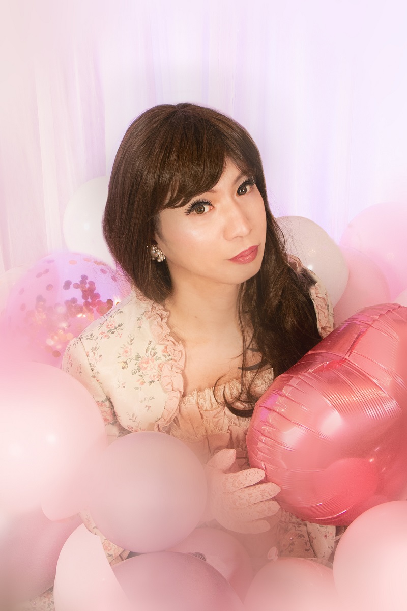 ピンクのバルーンに囲まれた空間で、花柄ドレスとレースのグローブに身を包んだ女装男子が微笑む姿を、プロのカメラマンが丁寧に撮影。フォトレタッチ済みの写真で、撮影場所は秘密の女装サロン「ベルマリ」。